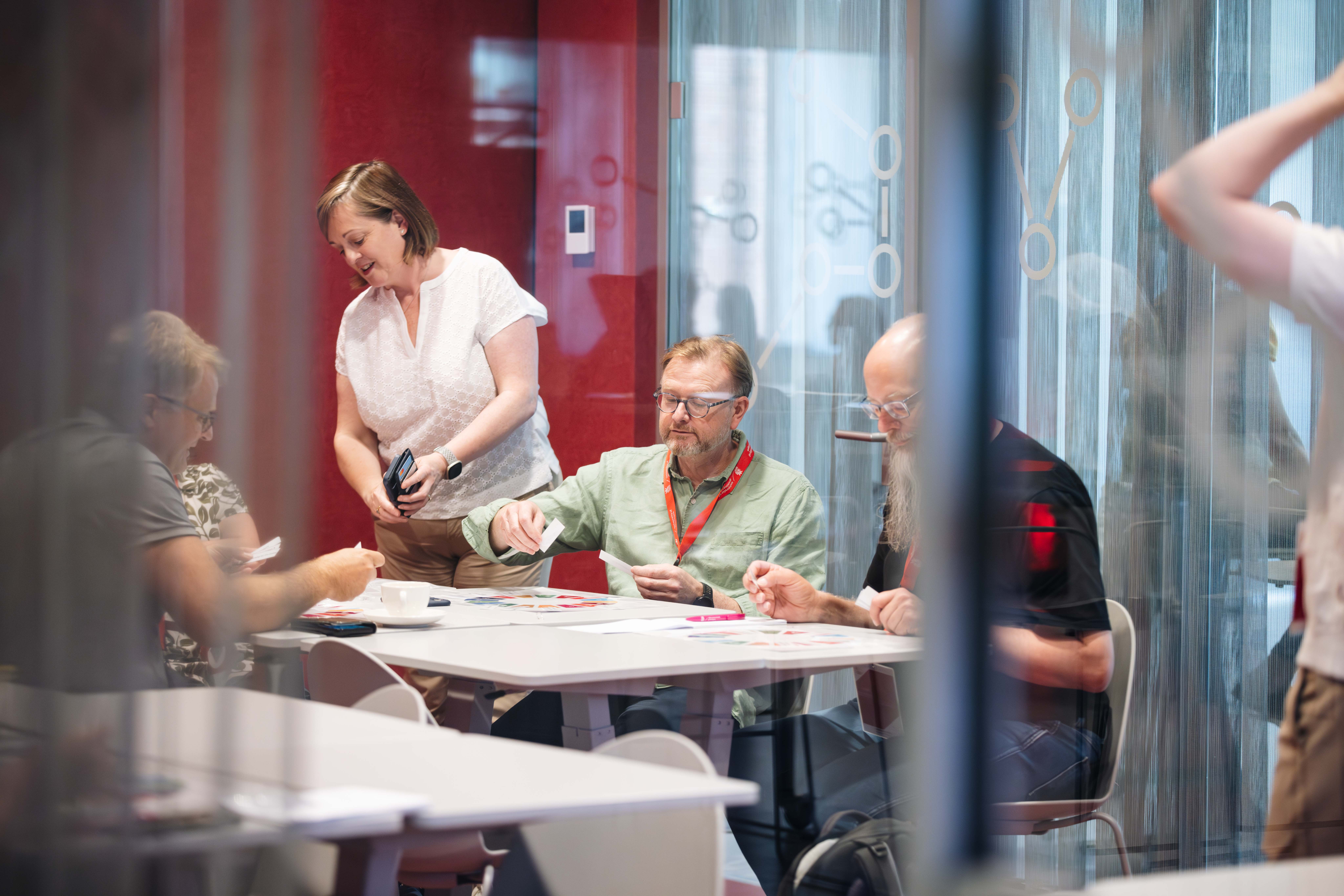 A group of people engaging in a collaborative activity at a round table, with a woman assisting the participants. The setting features a modern interior with glass walls and a red backdrop.